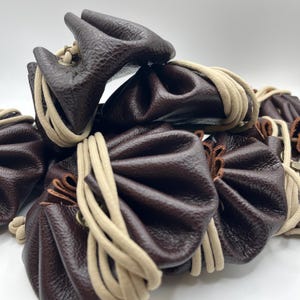 Pode incluir: Uma pilha de pequenas bolsas de couro marrom escuro com uma superfície texturizada. Cada bolsa é reunida no topo e presa com um cordão bege. As bolsas estão empilhadas, criando um efeito texturizado e em camadas.