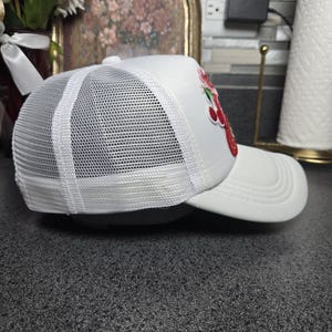 Op de afbeelding: Witte truckerpet met een mesh achterkant en een gebogen klep. De pet heeft een rood en groen geborduurd aardbei-ontwerp op de voorkant. Een klassieke stijl, geschikt voor vrijetijdskleding.