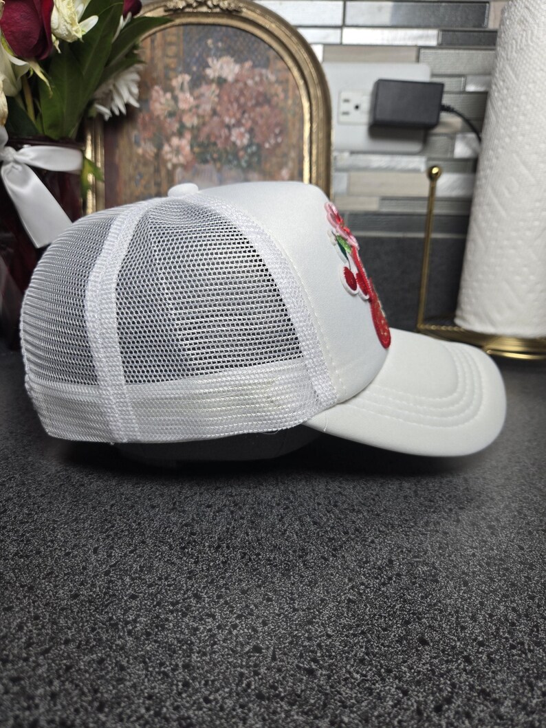 Op de afbeelding: Witte truckerpet met een mesh achterkant en een gebogen klep. De pet heeft een rood en groen geborduurd aardbei-ontwerp op de voorkant. Een klassieke stijl, geschikt voor vrijetijdskleding.