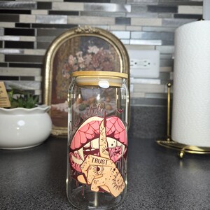 Peut inclure: Un gobelet en verre transparent en forme de canette avec un couvercle en bambou. Le gobelet pr&eacute;sente un graphisme de l&egrave;vres roses, une main faisant un doigt d'honneur et le mot "TRUST". Le design a une esth&eacute;tique de style tatouage.