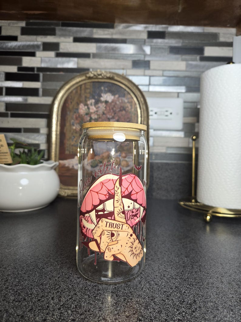 Peut inclure: Un gobelet en verre transparent en forme de canette avec un couvercle en bambou. Le gobelet pr&eacute;sente un graphisme de l&egrave;vres roses, une main faisant un doigt d'honneur et le mot "TRUST". Le design a une esth&eacute;tique de style tatouage.