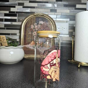 Peut inclure: Une canette &agrave; boisson en verre transparent avec un couvercle en bambou. La canette pr&eacute;sente un graphisme de l&egrave;vres roses, une main avec un doigt sur les l&egrave;vres et le mot "TRUST". Les mots "Basic Witch" sont au-dessus des l&egrave;vres.
