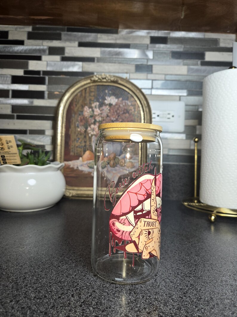 Peut inclure: Une canette &agrave; boisson en verre transparent avec un couvercle en bambou. La canette pr&eacute;sente un graphisme de l&egrave;vres roses, une main avec un doigt sur les l&egrave;vres et le mot "TRUST". Les mots "Basic Witch" sont au-dessus des l&egrave;vres.