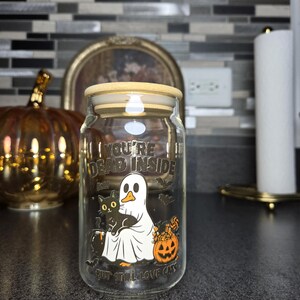 Może przedstawiać: Przezroczysty szklany słoik z drewnianą pokrywką z motywem Halloween. Słoik ma napis "YOU'RE DEAD INSIDE BUT STILL LOVE CATS" oraz grafikę ducha, czarnego kota i dyni. Słoik stoi na ciemnej powierzchni.