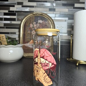Peut inclure: Un gobelet en verre transparent en forme de canette avec un couvercle en bambou. Le gobelet pr&eacute;sente un dessin d'une bouche, d'une main et d'un soleil avec le mot "TRUST". De petites illustrations de chauves-souris sont &eacute;galement pr&eacute;sentes.