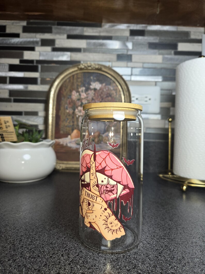 Peut inclure: Un gobelet en verre transparent en forme de canette avec un couvercle en bambou. Le gobelet pr&eacute;sente un dessin d'une bouche, d'une main et d'un soleil avec le mot "TRUST". De petites illustrations de chauves-souris sont &eacute;galement pr&eacute;sentes.