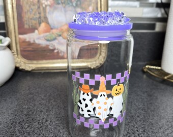 Peut inclure: Un bocal en verre transparent avec un couvercle violet et un motif décoratif représentant trois fantômes de dessins animés avec des chapeaux et une citrouille. Le bocal a une bordure violette à carreaux.
