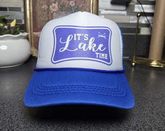 May include: A blue and white trucker hat with the text "IT'S Lake TIME" in a blue rectangle. The hat has a blue brim and a white front panel with a blue graphic. The hat is designed for outdoor activities.