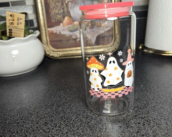 May include: A clear glass tumbler with a coral-colored lid. The tumbler features a whimsical design of three ghosts, a mushroom, and a pumpkin, all adorned with floral accents. The design is set against a black and pink checkered pattern.