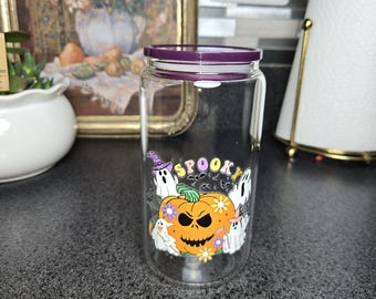 May include: A clear glass jar with a purple lid, featuring a Halloween-themed design. The design includes a jack-o'-lantern, ghosts, and the word "Spooky" in colorful letters. The jar is designed to hold beverages.