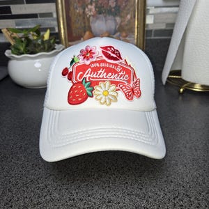 Peut inclure: Casquette de camionneur blanche avec une visière incurvée. Le chapeau présente un écusson avec des fleurs brodées, une fraise, un papillon et les mots "100% Original & Authentic". Il a un dos en maille blanche.