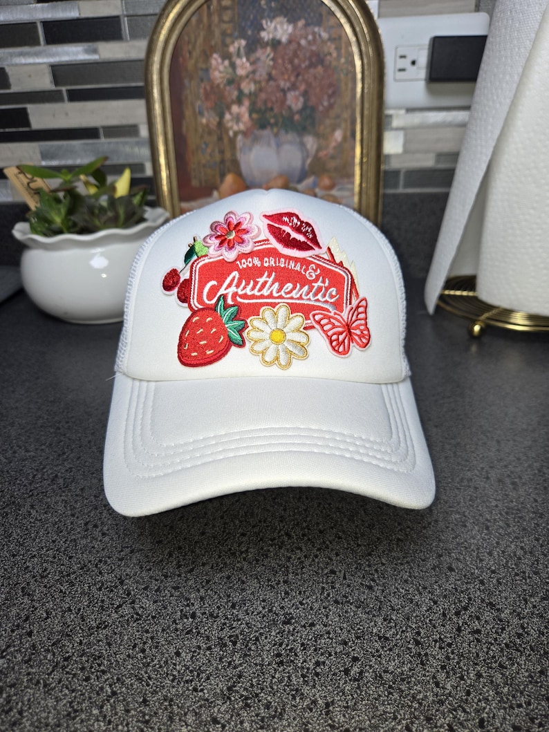 Op de afbeelding: Witte truckerpet met een gebogen klep. De pet heeft een patch met geborduurde bloemen, een aardbei, een vlinder en de woorden "100% Original & Authentic". Het heeft een witte mesh achterkant.