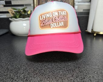 Può includere: Cappellino da camionista rosa con pannello frontale bianco e visiera rosa. Il cappello presenta il testo "Living on that Blue Collar Dollar" in un design marrone e arancione.