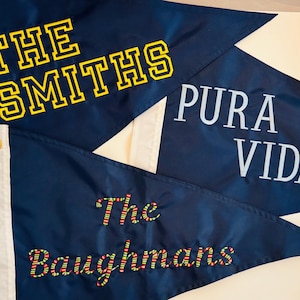 May include: Three triangular navy blue pennants with white side headers and metal grommets. One features yellow block text reading THE SMITHS, another has white text reading PURA VIDA, and the third displays The Baughmans in multicolored stitched lettering.