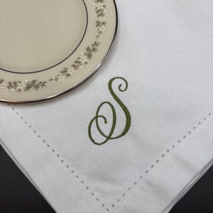 May include: A white linen napkin with a decorative green embroidered letter "S" and a decorative stitched border. A portion of a cream-colored plate with a floral design is in the upper left corner.