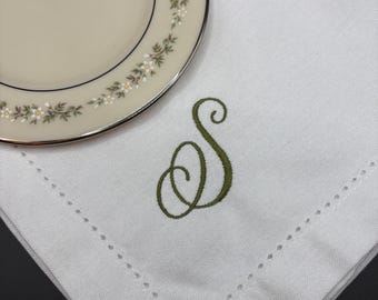 May include: A white linen napkin with a decorative green embroidered letter "S" and a decorative stitched border. A portion of a cream-colored plate with a floral design is in the upper left corner.