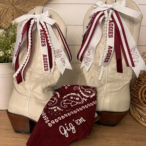 May include: A pair of off-white cowboy boots decorated with maroon and white ribbons featuring the text "AGGIES" and "gig 'em" alongside a maroon bandana with a white paisley pattern and the embroidered text "gig 'em" placed in front.