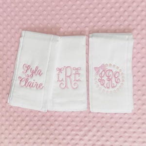 May include: Three white baby burp cloths with pink embroidered designs. One cloth features the name "Lyla Claire", another has a monogram, and the third has a monogram within a circle with a bow. The cloths are on a pink, textured surface.