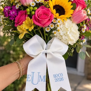 May include: A vibrant floral bouquet featuring pink roses, a yellow sunflower, pink tulips, white hydrangeas, and purple stock flowers. A white satin ribbon is tied around the stems, embroidered with blue text reading EUM and HOCO 2026.