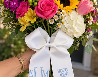 May include: A vibrant floral bouquet featuring pink roses, a yellow sunflower, pink tulips, white hydrangeas, and purple stock flowers. A white satin ribbon is tied around the stems, embroidered with blue text reading EUM and HOCO 2026.