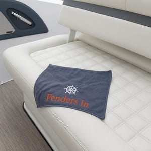 May include: A gray boat towel with the words "Fenders In" in orange and a white ship's wheel design. The towel is placed on a white quilted boat seat. The boat interior also features a dark blue panel.