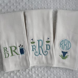 May include: Three white golf towels with embroidered designs. Each towel features a monogram and golf-themed elements in green and blue. One towel has a golf bag, another has a golf flag, and the third has a golf ball on a tee.