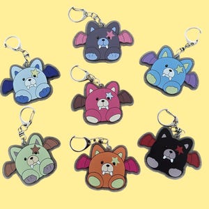 Op de afbeelding: Een verzameling kleurrijke sleutelhangers in de vorm van beren. Elke sleutelhanger heeft een beer met vleermuisvleugels, een ster en hoektanden. De sleutelhangers zijn in verschillende kleuren, waaronder blauw, roze, groen, oranje, zwart en paars, met zilveren sleutelringen.