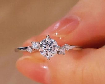 May include: A silver ring with a large, round, clear gemstone as the focal point. Smaller, clear gemstones are set on either side of the main stone, creating a cluster design. The ring is held against a blurred background.