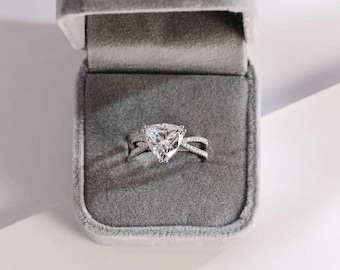 Pode incluir: Um anel de prata com uma grande pedra preciosa transparente triangular. O anel tem um design de banda cruzada e é exibido em uma caixa de anel de veludo cinza aberta. É uma joia.