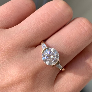 May include: A gold ring with a large, round, clear gemstone as the focal point. The ring features two smaller, rectangular gemstones on either side of the main stone. The ring is set in a gold band.