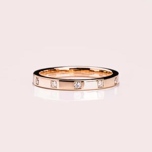 Op de afbeelding: Een roségouden bandring met vierkante diamanten gelijkmatig rond de band gezet. De ring heeft een gepolijste afwerking en wordt gepresenteerd op een lichtroze oppervlak. Dit is een sieraad.