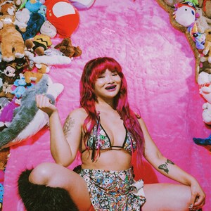 May include: A person wearing a patterned bikini top and skirt, with black fur leg warmers, sitting on a pink surface. The background is filled with stuffed animals. The person has pink hair.