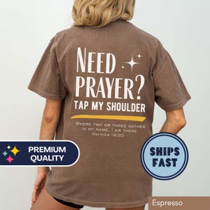 May include: Espresso-colored t-shirt with the text "Need Prayer? Tap My Shoulder" in white. Below the text is a yellow banner with the scripture Matthew 18:20. The shirt also has a "Premium Quality" and "Ships Fast" badge.