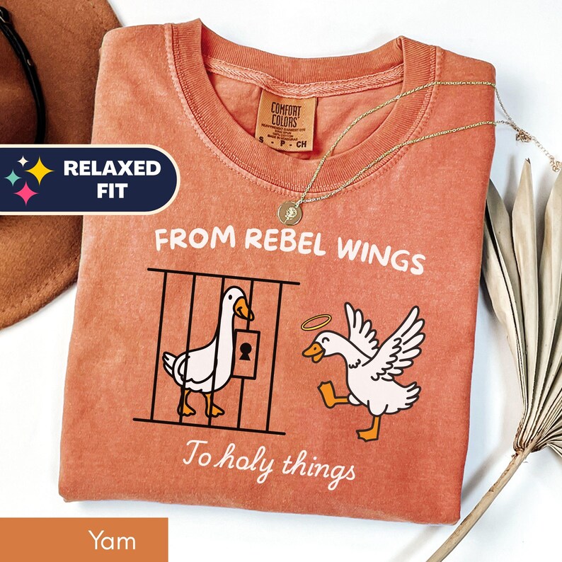 May include: A yam-colored t-shirt with the text "FROM REBEL WINGS To holy things" and a graphic of two geese. One goose is in a cage, and the other has a halo. A gold necklace is draped on the shirt. The shirt has a relaxed fit.