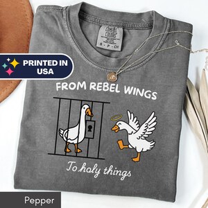 May include: Pepper-colored t-shirt with a graphic of two geese, one in a cage and the other with a halo. Text reads "FROM REBEL WINGS To holy things" and "PRINTED IN USA". A Comfort Colors tag is visible.