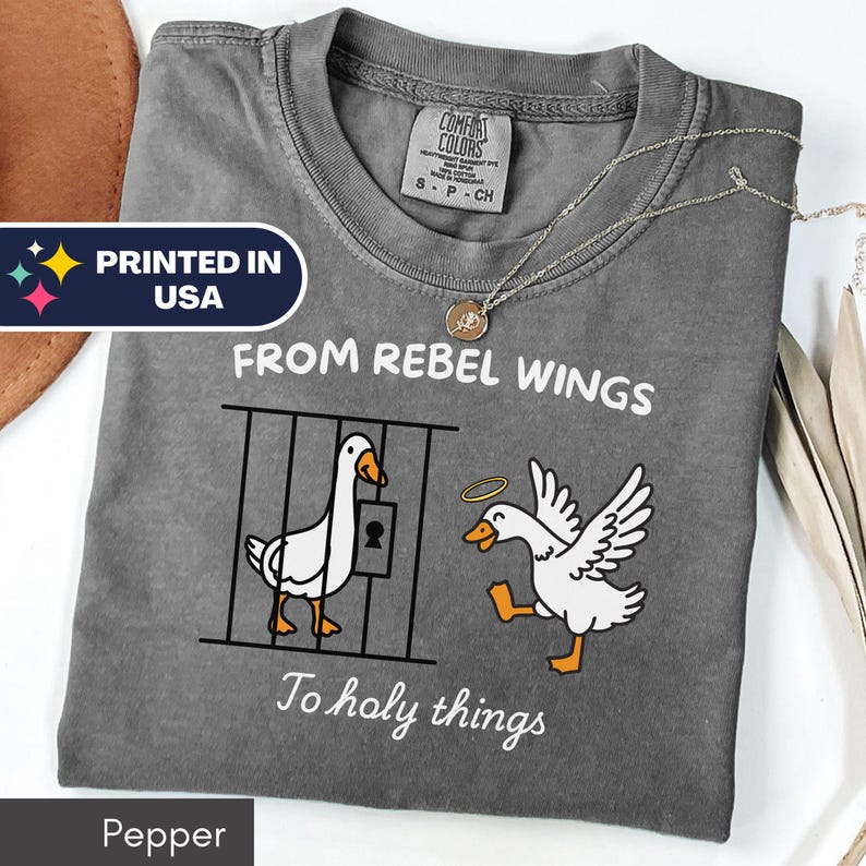 May include: Pepper-colored t-shirt with a graphic of two geese, one in a cage and the other with a halo. Text reads "FROM REBEL WINGS To holy things" and "PRINTED IN USA". A Comfort Colors tag is visible.