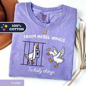 May include: Violet-colored t-shirt with the text "FROM REBEL WINGS To holy things" and a graphic of two geese. One goose is behind bars, and the other has a halo. The shirt is labeled "100% Cotton."