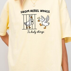 May include: A light yellow t-shirt with a graphic of two cartoon geese. One goose is behind bars, and the other has a halo and wings. Text on the shirt reads "FROM REBEL WINGS To holy things". The word "Banana" is at the bottom.