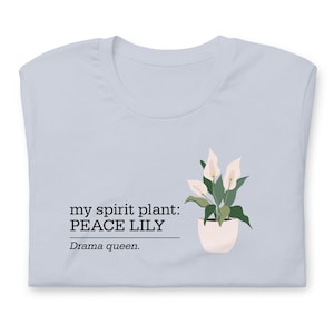 Puede incluir: Camiseta azul claro con un gráfico de una flor de lirio de la paz en una maceta blanca. El texto "my spirit plant: PEACE LILY" está encima de la planta, con "Drama queen." debajo.
