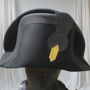 May include: A black bicorne hat with a gold emblem and black trim. The hat is displayed on a black mannequin head. The hat has a wide brim and a decorative black fan-shaped element.