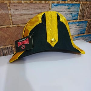 Peut inclure: Un chapeau bicorne vert foncé avec une garniture dorée et un bouton doré. Le chapeau a une bande noire et une étiquette qui dit "EURO ERA BUSINESS CO."