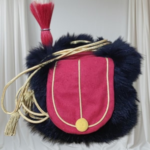Può includere: Una borsa in stile militare con una tasca rosso scuro, finiture dorate e una chiusura con bottone dorato. La borsa è circondata da pelliccia blu scuro e ha una corda e un nappa dorate. Una piuma rossa e bianca è attaccata in alto.