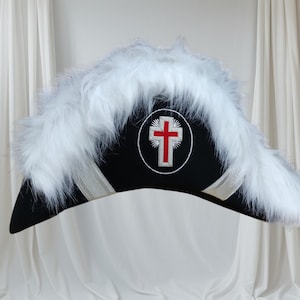 May include: A black hat with white fur trim and a red cross emblem. The hat has a triangular shape with a curved brim and is decorated with white ribbon accents. The background is white fabric.