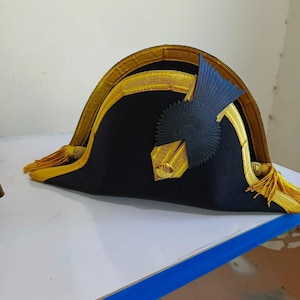 May include: A black bicorne hat with gold trim and tassels. The hat features a dark blue emblem on the front. The hat is likely a historical or costume piece.