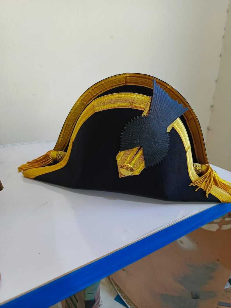 May include: A black bicorne hat with gold trim and tassels. The hat features a dark blue emblem on the front. The hat is likely a historical or costume piece.