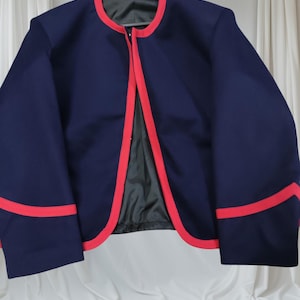 May include: A navy blue jacket with red trim around the collar, front edges, and sleeve cuffs. The jacket is hanging on a black hanger against a white background. The jacket has a black lining.