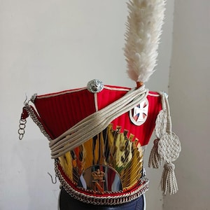 Può includere: Un cappello militare rosso con dettagli dorati e argentati, una piuma bianca e un bordo nero. Il cappello presenta un'emblema dorato, una catena d'argento e una croce bianca. Stile militare storico.
