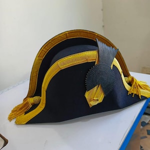 May include: A dark blue bicorne hat with gold trim and tassels. The hat has a decorative black emblem and gold detailing. This hat is likely a historical or theatrical costume piece, reminiscent of naval attire.