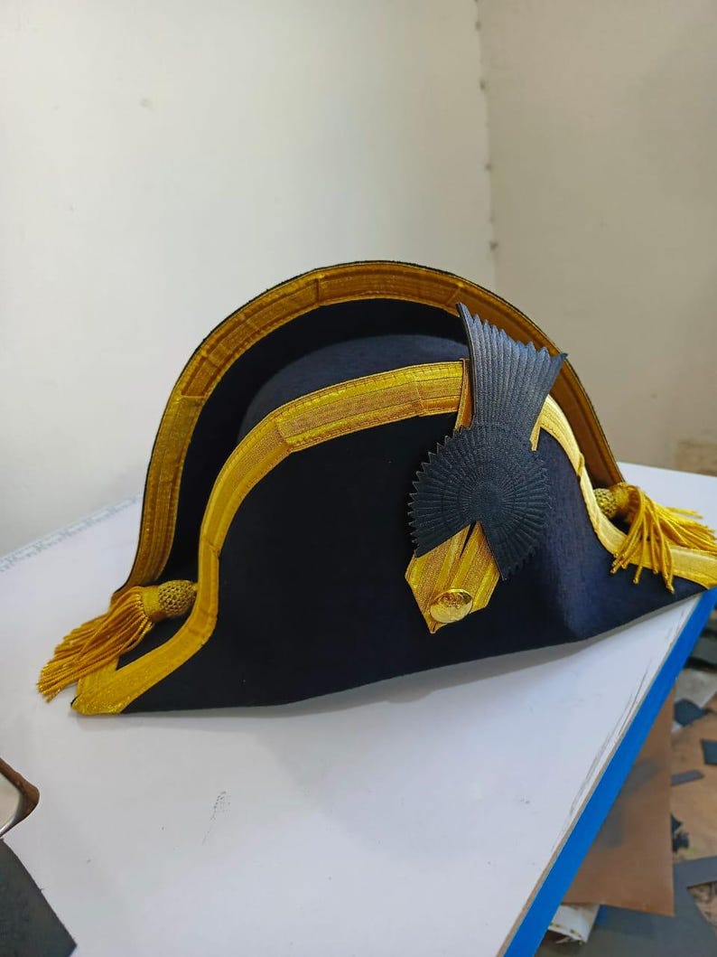 May include: A dark blue bicorne hat with gold trim and tassels. The hat has a decorative black emblem and gold detailing. This hat is likely a historical or theatrical costume piece, reminiscent of naval attire.