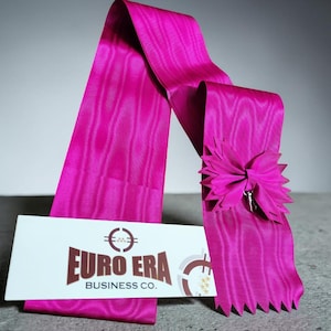 Peut inclure: Un ruban fuchsia vif avec une rosette décorative et un petit charme argenté. Le ruban est drapé sur une carte blanche avec le texte "EURO ERA BUSINESS CO." en marron. Le ruban a un aspect texturé.
